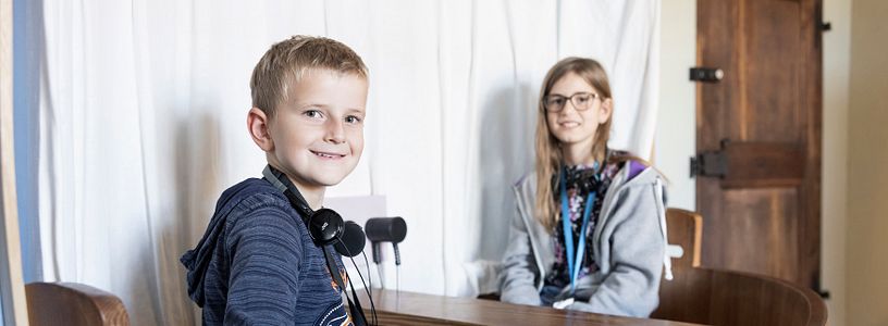 Ein Bub und ein Mädchen sitzen mit Kopfhörern in einem kleinen Schiff in der Ausstellung von Schloss Hallwyl. Der Bub blickt direkt in die Kamera.