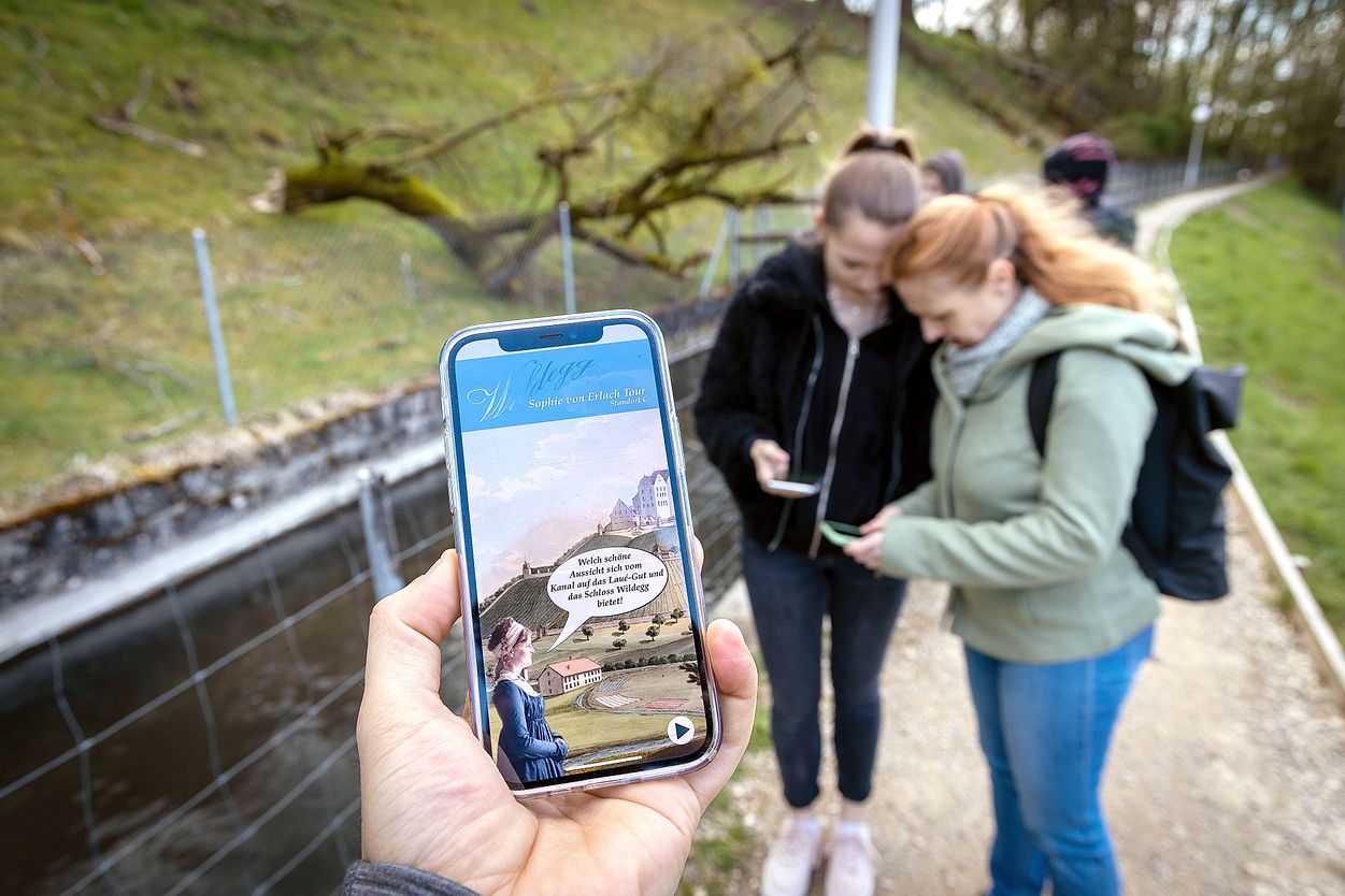 Nahaufnahme eines Mobiltelefons. Auf dem Screen ist die App "Industriekultur Aabach" sichtbar. Im Hintergrund sind mehrere Personen in Unschärfe zu sehen.