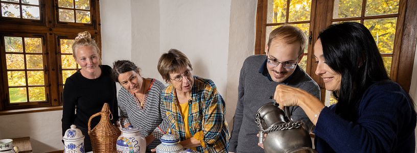 Eine Besuchergruppe in einem historischen Raum von Schloss Hallwyl. Die Gruppe steht um einen Tisch herum, auf dem Gefässe stehen. Eine Frau geisst eine Flüssigkeit aus einem Metallkrug in einen Metallbecher.