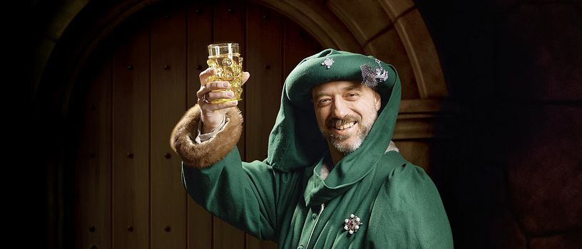 Ein mittelalterlich gekleideter Mann. Er blickt direkt in die Kamera und hält feiernd ein Glas mit Bier in die Höhe.