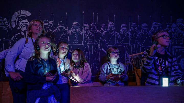 Eine Familie in einem dunklen Raum; die Displays der Audiogeräte um den Hals leuchten mystisch im Dunkeln. An der Rückwand Silhouetten von Legionären.
