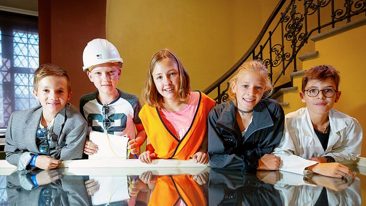 Kinder als Forscher und Forscherinnen gekleidet lehnen sich an einen spiegelnden Tisch