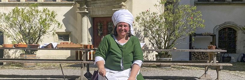 Eine mittelalterlich gekleidete Frau sitzt an einem Feuer im Hof von Schloss Lenzburg und kocht. 