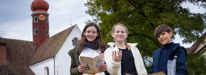 Zwei Mädchen und ein Bub stehen gemeinsam vor dem Kloster Wettingen und blicken direkt in die Kamera. Sie halten eine Landkarte, Broschüre sowie eine Pyramide in den Händen.