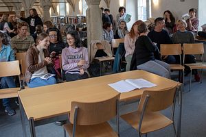 Ganz vorne ist ein Tisch mit zwei Stühlen gegen ein Publikum gerichtet, das aus mehreren Jugendlichen und Erwachsenen besteht. Gewisse Personen stehen im Hintergrund noch. Dahinter sind Bücherregale zu sehen, es handelt sich um eine Bibliothek. In der Mitte des Raumes gibt es vier Steinsäulen, die sich von vorne bis hinten ziehen.