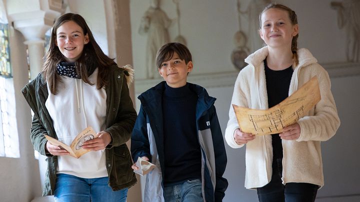 Zwei Mädchen und ein Bub schreiten nebeneinander durch den Kreuzgang des Klosters Wettingen, sie blicken lachend in die Kamera.