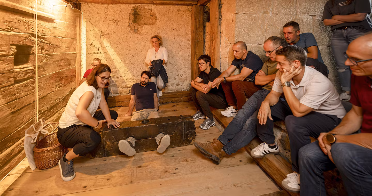 Eine Gruppe Männer und Frauen sitzen mit einer Museumsführerin im Gefängnis von Schloss Lenzburg. Eine Person liegt am Boden, die Füsse sind in einem Holz fixiert