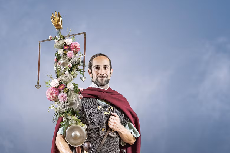 Ein römischer Legionär vor blauem Hintergrund. Er trägt eine mit Blumen geschmückte Standarte in der rechten Hand. 