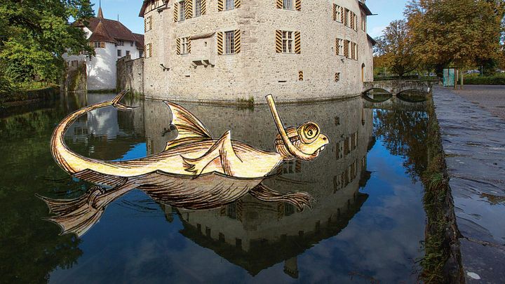 Aussenaufnahme von Schloss Hallwyl mit Aabach im Vordergrund. Im Wasser schwimmt ein Drachentier mit Schnorchel (Illustration), am Himmel ist ein Luftschiff zu sehen (Illustration)