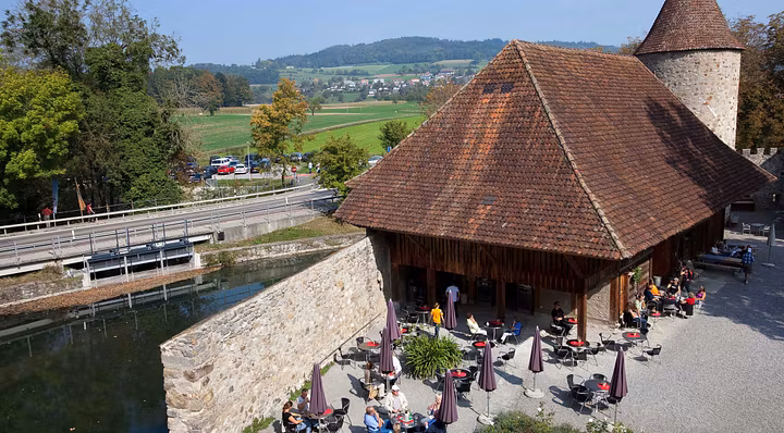 Blick von oben auf den Aussenbereich des Cafés von Schloss Hallwyl. An den Tischen im Schlosshof sitzen Besucherinnen und Besucher. Das Wetter ist sommerlich.