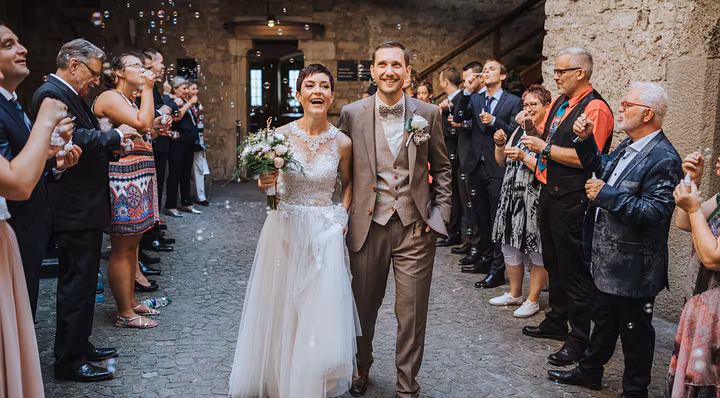 Ein Hochzeitspaar auf Schloss Habsburg: Ein Mann und eine Frau schreitet durch spalierstehende Gäste. Seifenblasen fliegen in der Luft.
