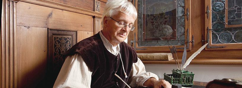 Ein mittelalterlich gekleideter Mann (Kaligraph) schreibt mit Feder und Tinte auf Schloss Lenzburg.