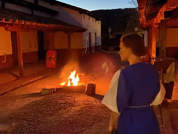 Eine römisch gekleidete Frau steht in der Gasse der Mannschaftsunterkunft im Legionärspfad Vindonissa. Es ist Abend und das Licht einer Feuerschale erhellt die Umgebung.