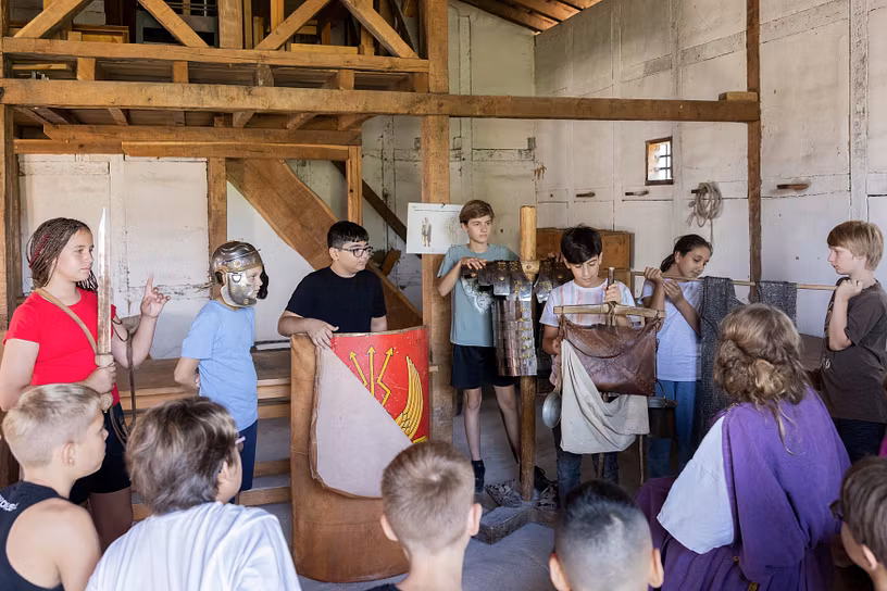 Eine Schulklasse steht in der Mannschaftsunterkunft (Contubernia) auf dem Legionärspfad Vindonissa und betrachtet unterschiedliche Ausrüstungsgegenstände eines Legionärs. 