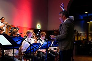 Im Bild sind links mehrere musizierende Personen zu sehen. Alle spielen verschiedene Blasinstrumente und haben ein weisses Hemd an. Vor den sitzenden Bandmitglieder steht der Dirigent in einem grauen Jackett.  