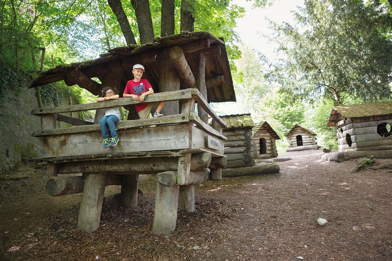 Spielplatz Schloss Lenzburg: Mehrere Holzhäuschen. Auf dem vordersten Häuschen stehen mehrere Kinder und blicken direkt in die Kamera.