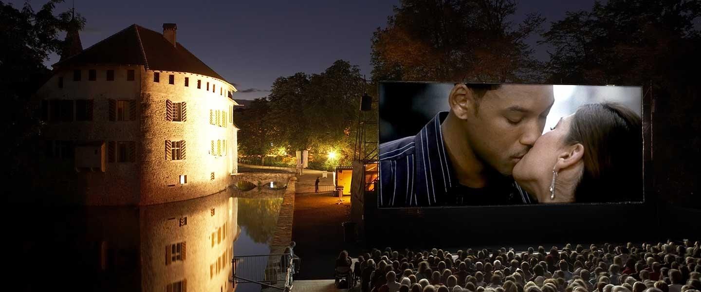 Nachtaufnahme von Schloss Hallwyl mit Aabach, rechts eine grosse Leinwand mit einer Filmszene (ein küssendes Paar).