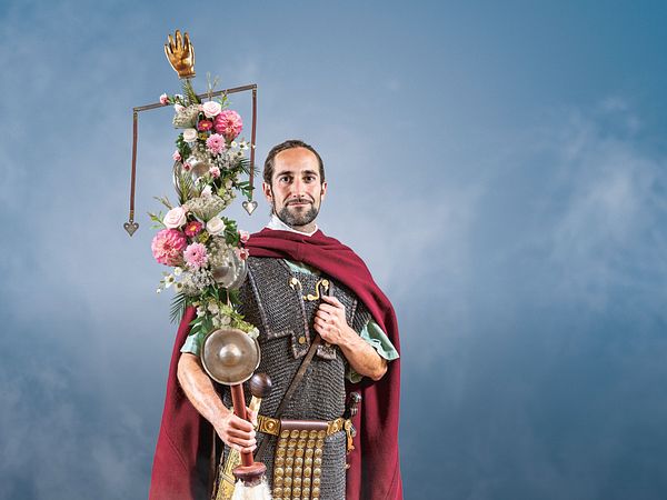 Ein römischer Legionär mit Standarte (geschmückt mit Blumen) vor blauem Hintergrund.