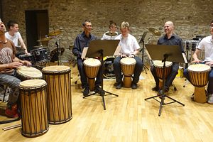 Mehrere Personen in einem Halbkreis spielen gemeinsam auf Trommeln Musik. Im Hintergrund spielen zwei Personen auf dem Schlagzeug.