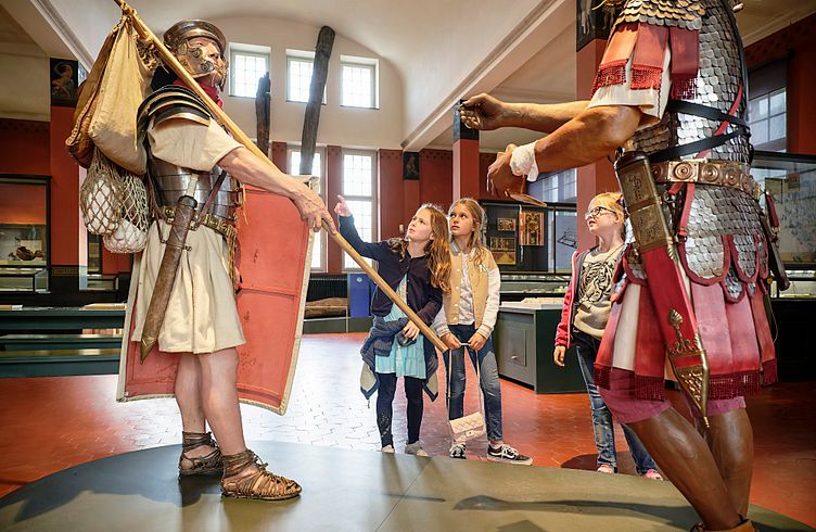Drei Mädchen stehen in der Ausstellung im Vindonissa Museum. Vor ihnen stehen zwei lebensgrosse Legionäre (Figuren)