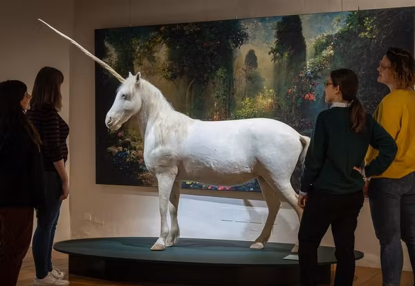 Vier Menschen stehen in einer Ausstellung um ein präpariertes Einhorn. Hinter dem Einhorn ist ein grosses Bild von einem verwunschenen Wald zu sehen.