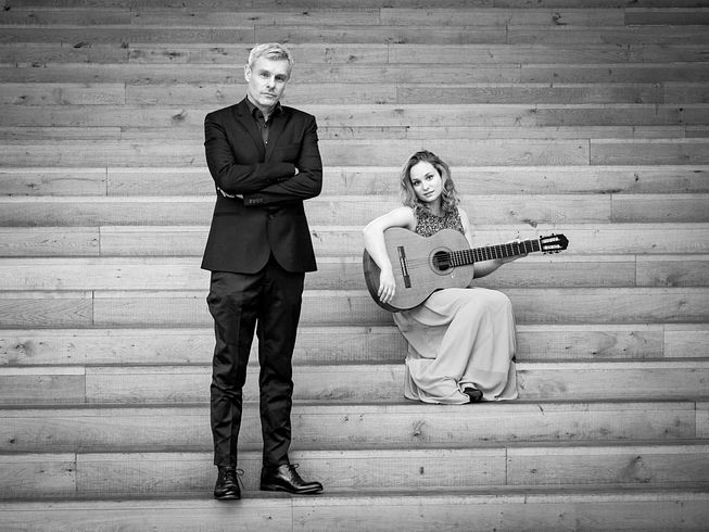 Schwarz-weiss-Aufnahme von "The Art of the Duo": Ein Mann steht, eine Frau dahinter sitzt mit einer Gitarre auf einer Treppe. 