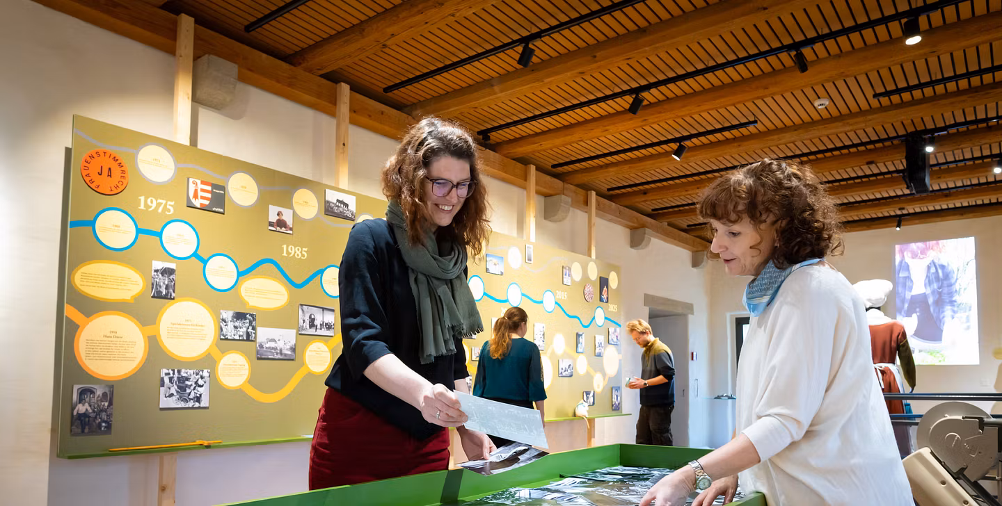 Ausstellungsraum im Schloss Lenzburg: Zwei Frauen stehen an einem Tisch und stöbern in alten Fotos, im Hintergrund weitere Ausstellungselemente.