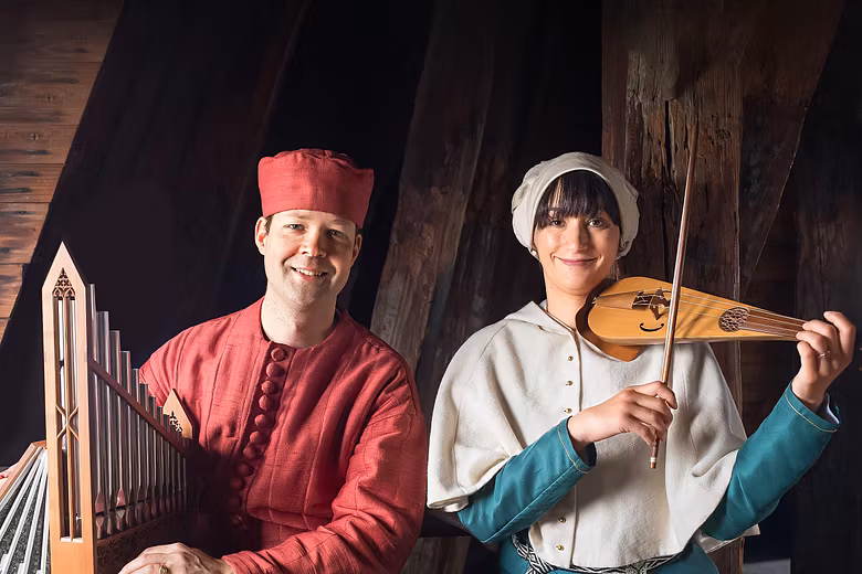 Ein mittelalterlich gekleideter Musiker neben einer mittelalterlich gekleideten Musikerin. Im Hintergrund ein Raum aus Holz.