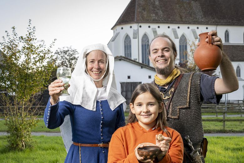 Eine mittelalterlich gekleidete Frau, ein römischer Legionär sowie ein modern gekleidetes Mädchen. Sie blicken alle freudig in die Kamera.