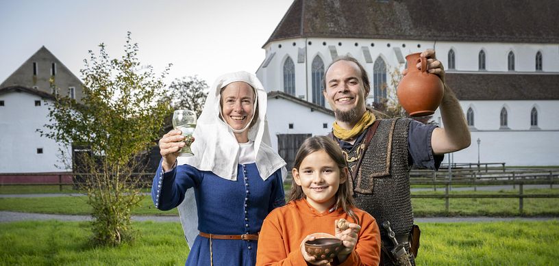 Eine mittelalterlich gekleidete Frau, ein römischer Legionär sowie ein modern gekleidetes Mädchen. Sie blicken alle freudig in die Kamera.
