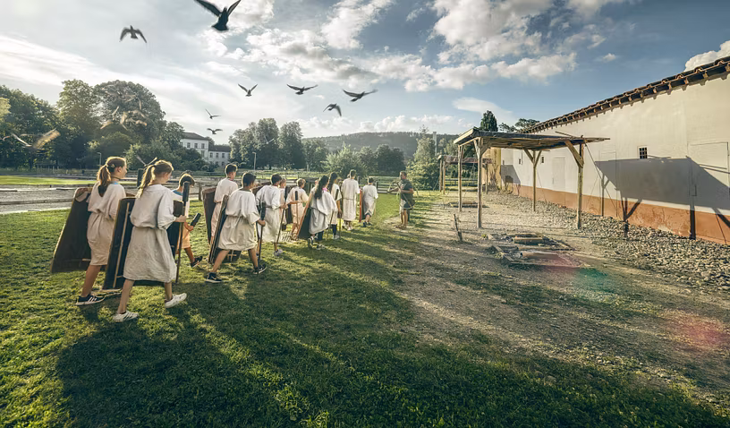 Eine Gruppe Schülerinnen und Schüler marschiert in römischer Tunika wie römische Legionäre. Darüber ein Himmel mit Vögeln.