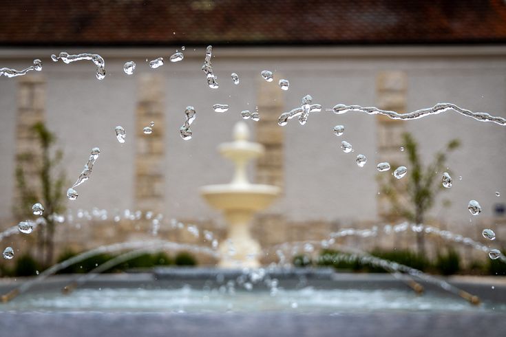 Nahaufnahme von Wassertropfen (eingefroren durch Kurzzeitbelichtung). Im Hintergrund ein antiker Brunnen im römischen Garten vom Vindonissa Museum.