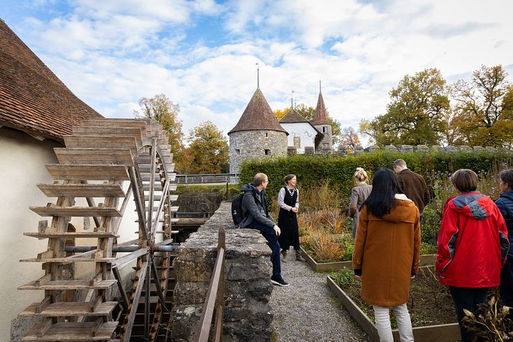 Eine Gästegruppe und eine Museumsführerin steht neben der Mühle von Schloss Hallwyl. Links ein grosses Mühlenrad, im Hintergrund Schloss Hallwyl