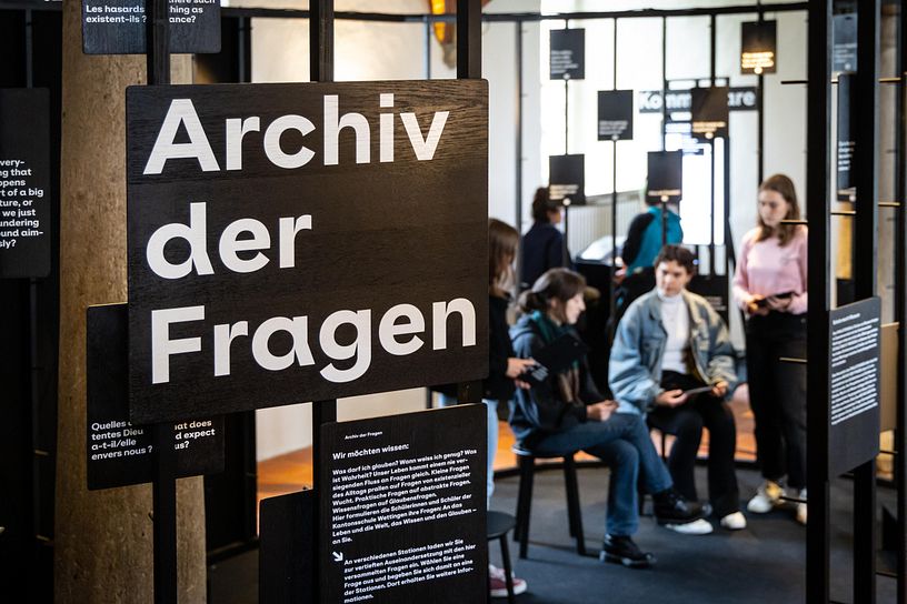 Ausstellungsraum im Parlatorium der Klosterhalbinsel Wettingen: Ein grosses Schild mit dem Text "Archiv der Fragen", im Hintergrund sind mehrere Personen zu sehen, die diskutieren.