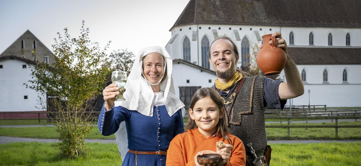 Eine mittelalterlich gekleidete Frau, ein römischer Legionär sowie ein modern gekleidetes Mädchen. Sie blicken alle freudig in die Kamera.