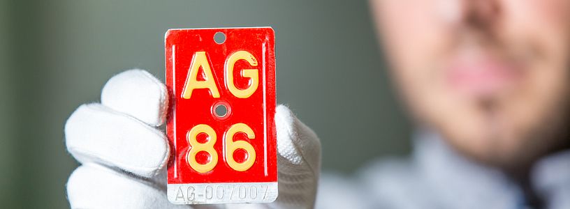 Rote Velonummer mit der Aufschrift "AG 86" in der Hand eines Mannes mit weissen Handschuhen. Im Hintergrund in Unschärfe der Kopf des Mannes (er hält die Velonummer in die Kamera).