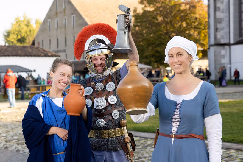 Gruppenbild mit einer Römerin, einem römischen Legionär und einer mittelalterlich gekleideten Magd. Die drei blicken in die Kamera und haltend prostens Weinkrüge in die Höhe.