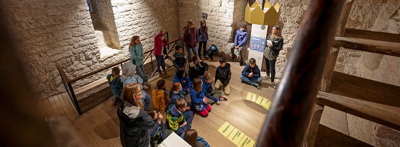 Eine Schulklasse sitzt im Kreis im Innern von Schloss Habsburg; am Boden liegen Blätter mit Texten.