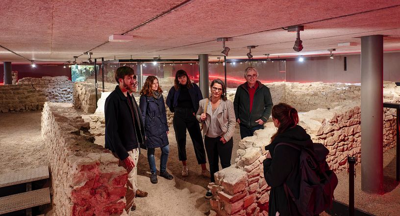 Besucherinnen und Besucher mit Museumsführerin in der versunkenen Offziersküche von Vindonissa. Die Gruppe steht inmitten von Steinmauern.