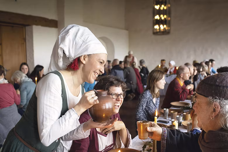 Gäste sitzen im kleinen Rittersaal von Schloss Lenzburg an langen Tischen und essen gemeinsam. Eine Frau in mittelalterlicher Kleidung giesst Getränke nach.