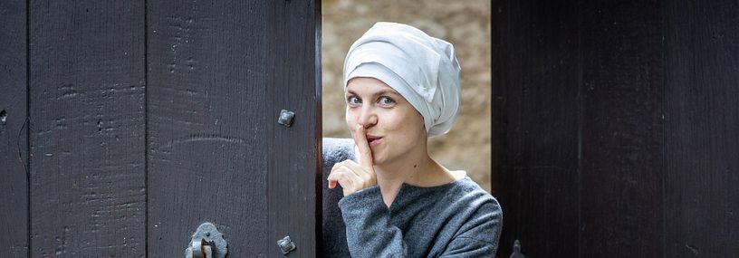 Eine mittelalterlich gekleidete Magd blickt durch das offene Tor von Schloss Habsburg in die Kamera. Sie hält verschwörerisch den linken Zeigfinger vor dem Mund ("nichts verraten!")
