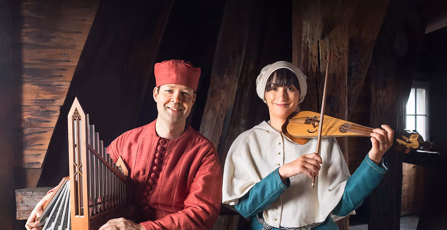 Ein mittelalterlich gekleideter Musiker neben einer mittelalterlich gekleideten Musikerin. Im Hintergrund ein Raum aus Holz.