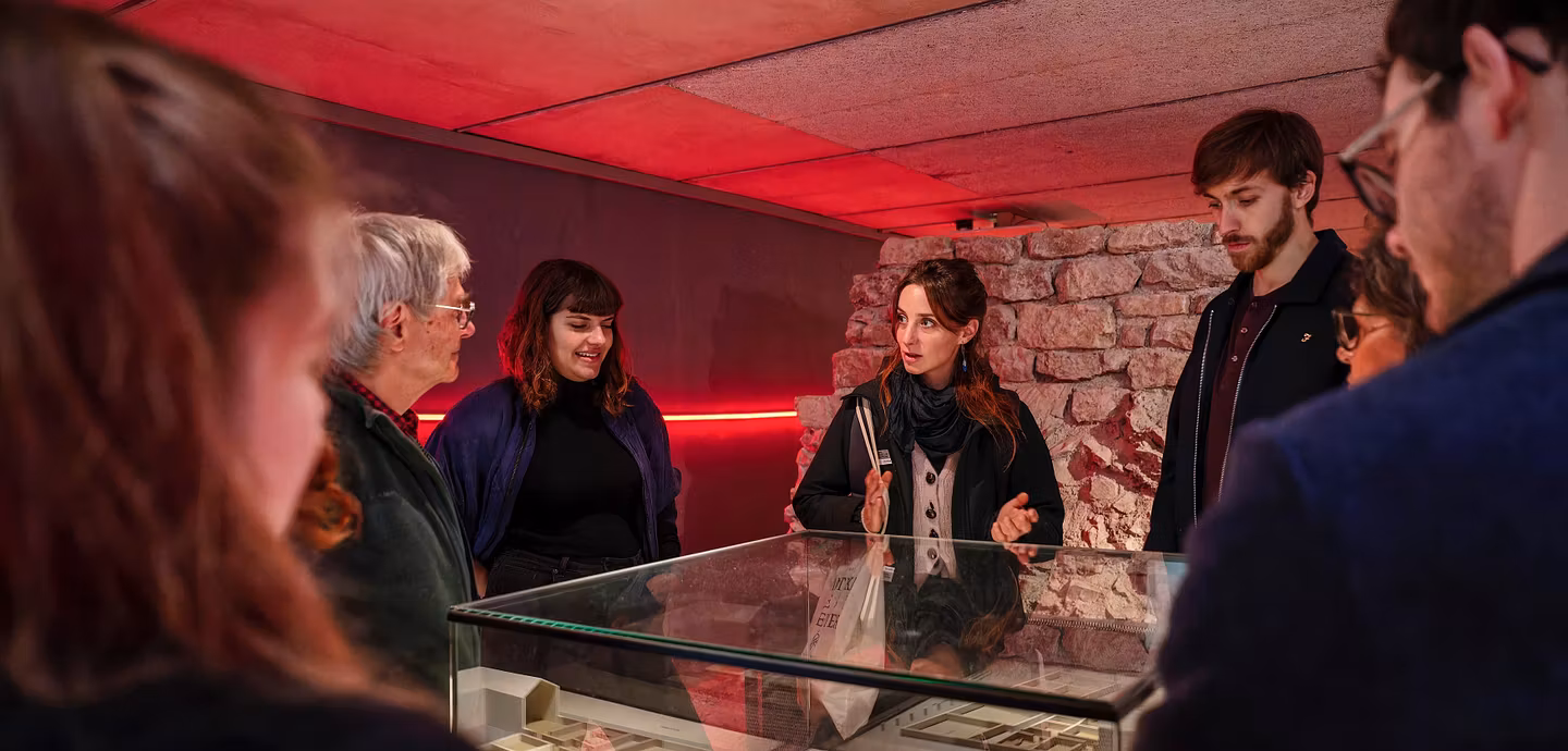 Eine Museumsführerin steht mit Besucherinnen und Besuchern um eine Vitrine in der römischen Offiziersküche im Legionärspfad Vindonissa. Im Hintergrund sind alte Mauerreste zu sehen.