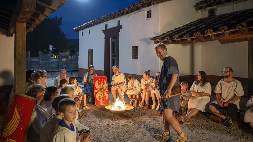 Eine grosse Gruppe mit Erwachsenen und Kindern. Sie sitzen um ein Lagerfeuer im Legionärspfad Vindonissa, in der Mitte brennt ein grosser Feuer. Daneben steht ein römischer Legionär, der lacht. Mystische Stimmung in der Nacht.
